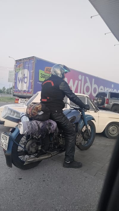 A side view of a person on a blue motorcycle is captured from a slightly elevated angle, possibly from within a vehicle. The rider is wearing black leather pants, a black vest with a red logo, and a silver helmet with blue accents. The motorcycle is laden with luggage, including rolled sleeping bags or blankets and a silver cylindrical object secured to the rear. In the background, a white car and a large blue truck with the "wildberries" logo are visible. A sign with Cyrillic text is also present. The scene appears to be on an asphalt road with paving stones bordering it, suggesting an urban or commercial area. The sky is overcast and hazy. The license plate on the motorcycle reads "4039 AA 19 RUS".
