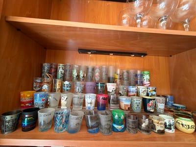 The image shows a collection of souvenir shot glasses displayed on two shelves inside a wooden cabinet. The top shelf is lined with wine glasses. The lower shelf contains approximately 50 shot glasses of various designs and sizes. Many of the shot glasses feature city names and landmarks, including Ávila, Barcelona, Benidorm, Cuenca, Holland, Amsterdam, Toledo, and Valencia. Some glasses appear to be clear with etched designs, while others are colorful and painted. A black LED light strip is affixed to the underside of the top shelf, illuminating the shot glasses. The cabinet's wooden finish is visible in the background. The location, Azuqueca de Henares, Spain, is not directly discernible from the image.