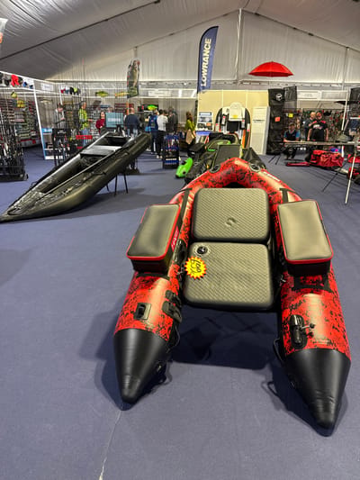The image depicts an indoor scene within a large white tent-like structure, identified as an exhibition space in the city of Mora, Portugal. The floor is covered with a blue carpet.

In the foreground, a red and black inflatable boat, possibly a belly boat or pontoon-style craft, is positioned centrally. It features a mottled red and black pattern on its inflatable tubes, black conical ends, and a grey textured seat with two dark grey side storage compartments. A yellow star-shaped price tag displaying "€99" and "€333" is affixed to the seat.

In the midground, to the left of the prominent red boat, a black inflatable kayak, possibly labeled "STAYCRAFT," rests on a dark metal display stand.

The background reveals multiple vendor booths. On the left, racks laden with numerous fishing rods and other angling equipment are visible. Towards the right, a blue banner displaying "LOWRANCE" hangs. Further right, a red umbrella is suspended, and more display areas are present with various products, including additional inflatable items and bags.

Several individuals are present throughout the background, walking, standing, and observing the displayed merchandise. Their actions are limited to browsing the exhibition. One individual in a yellow-green jacket is visible near the fishing rod display, and two men in black shirts are near the area with the red umbrella. The overall environment suggests an outdoor sports or fishing equipment trade show or exhibition.