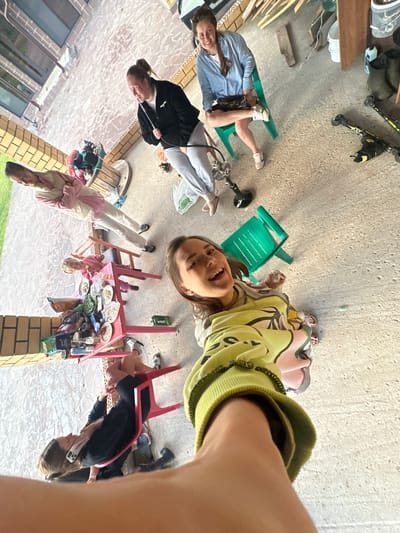 The image shows an outdoor scene at what appears to be a residential backyard or patio area. The ground is concrete, and there are brick elements visible on the left side, suggesting a building. There are several people present, including adults and a child. One woman is sitting on a green plastic chair, wearing a blue and white striped shirt and dark shorts, with her legs crossed. Another woman is standing, wearing a black jacket and grey pants, and holding a white object. A young child is sitting at a small red table covered with plates of food and various items. Another person is lying on a red chair, wearing black athletic shorts and a black long-sleeved shirt, with a phone in their hand. In the foreground, a person's arm and shoulder are visible, indicating the photo is taken from a first-person perspective. Several objects are scattered around, including skis, boots, a wooden spool, and plastic bags. The overall setting suggests a casual gathering or an informal outdoor activity. The image is taken in Пепеляева, Russia.