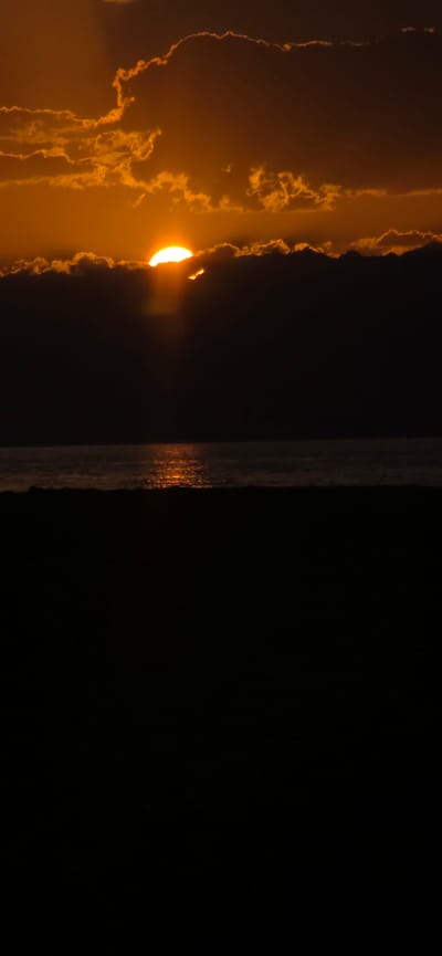 The image shows a sunset over the sea, with the sun partially obscured by dark clouds. The sky is filled with warm orange and yellow hues, and the sea reflects the sunset's glow. A dark silhouette of a beach or landmass is visible in the foreground. The scene appears peaceful and serene. This image was captured at the city of Basbeshik, Kyrgyzstan.