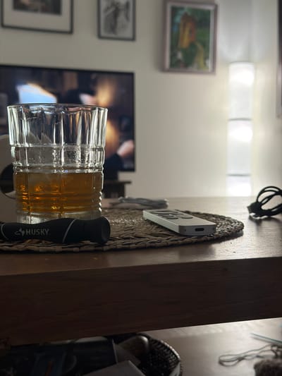 A partially filled whiskey glass and a Husky screwdriver lie on a wooden surface. In the background, there are two framed pictures on a wall, and a television screen displaying an image. The specific location is Buford, United States.