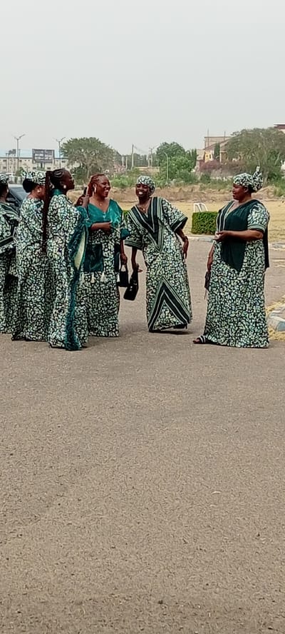 Six adult women are depicted outdoors on a paved asphalt surface, likely in Goron Dutse, Nigeria. They are gathered in a group, appearing to be engaged in animated conversation and laughter, conveying a sense of joy and camaraderie.
The women are uniformly dressed in long, loose-fitting garments, possibly traditional Nigerian kaftans or dresses, featuring a light green and white/cream floral or abstract pattern. Several of the outfits incorporate prominent darker green accents, particularly on the woman in the center and the two on the far right. Most of the women wear matching head coverings, completing their coordinated ensembles. Accessories include black handbags carried by at least three of the women, and sandals visible on the feet of the woman on the far right.
The background consists of sparse vegetation, including trees and bushes, and indistinct buildings under a hazy or overcast sky. Utility poles and lines are also visible, suggesting a semi-urban or urban fringe setting. The diffused light indicates daytime, possibly mid-morning or late afternoon, without strong shadows. No visible text is present in the image.