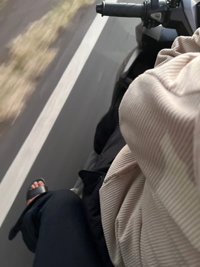 This is a close-up, low-angle shot taken from the perspective of someone riding a motorcycle. The primary subjects are the rider's legs and clothing, along with part of the motorcycle's handlebars. The rider is wearing black pants and black sandals, and a cream-colored corduroy jacket. The jacket has distinct horizontal ribbing.
The scene is a road, indicated by the grey asphalt and a white painted line running diagonally across the frame. The motion of the motorcycle is suggested by the blurred background of foliage and the road surface, implying the image was taken while moving.
The time of day and weather are not explicitly clear, but the lighting appears to be natural daylight. The overall mood is neutral, focusing on the sensory experience of riding. There is no visible text in the image. The location context of Purworejo, Indonesia, suggests a tropical or subtropical environment, though this is not directly evident from the visual information alone. The image captures a moment of travel, emphasizing the personal experience of being on a motorcycle.