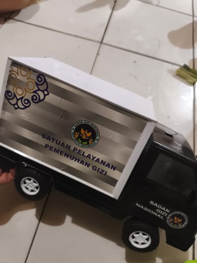 A black toy pickup truck with a white enclosed cargo bed is situated on a light-colored tiled floor with dark grout lines, observed indoors in the city of Cileungsi, Indonesia. The side of the cargo bed displays a prominent sticker featuring the circular logo of "BADAN GIZI NASIONAL REPUBLIK INDONESIA" (National Nutrition Agency of the Republic of Indonesia), above the text "SATUAN PELAYANAN PEMENUHAN GIZI" (Nutrition Fulfillment Service Unit). Decorative gold and blue swirling patterns adorn the upper left corner of the cargo bed. The truck's cab also bears the text "BADAN GIZI NASIONAL" and a smaller version of the agency's logo. The vehicle is equipped with black tires and white five-spoke rims. A human hand is partially visible on the left, near the front wheel. The background shows blurred floor tiles and an out-of-focus green object on the far right.