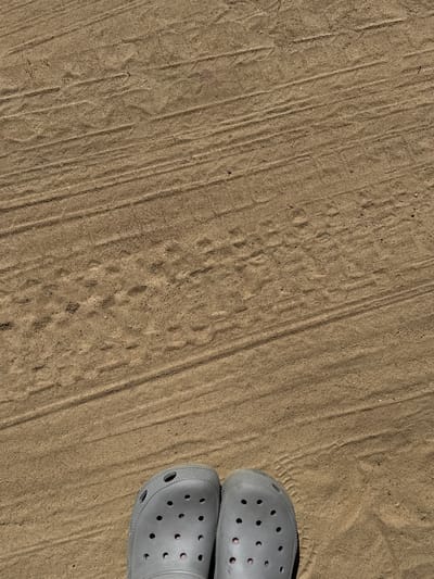 The image depicts a close-up, overhead view of two gray Crocs shoes standing on a sandy surface. The sand displays visible tire tracks and some faint impressions of foot or paw prints. The lighting suggests a bright, outdoor environment. The location is identified as Yevrostroy, Russia.