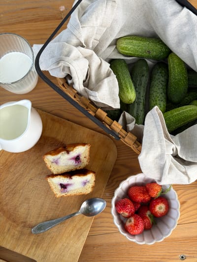 The image displays a top-down view of a wooden table with a collection of food items. In the upper left, a woven basket is filled with fresh cucumbers, partially covered by a light-colored cloth. To the right of the basket, a small, scalloped bowl holds several ripe strawberries. In the lower half of the image, a wooden cutting board features two slices of what appears to be a berry-filled cake, with a spoon resting nearby. A small white pitcher and a glass filled with milk are also present on the table, towards the bottom left. The setting appears to be indoors, given the lighting, and the wooden table suggests a rustic or home environment. There are no people visible, and the scene is static, focused on the presentation of the food. The overall impression is one of a simple, wholesome meal or snack preparation. The presence of fresh produce and cake suggests a casual meal or perhaps ingredients for a recipe. The lighting suggests it might be daytime.