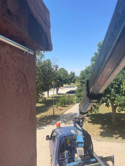 A blue utility truck with a crane attachment is parked on a paved area next to a building. The truck has an orange emergency light on its roof. A metal pipe is mounted to the exterior wall of the building. Trees and a street are visible in the background. The sky is clear and blue. This scene is located in the city of Zlatovrah, Bulgaria.