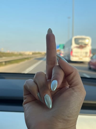 The image displays a human hand, appearing to be female, with a light skin tone, positioned centrally in the foreground. The hand is making an obscene gesture, with the middle finger extended vertically and the other fingers curled inwards. The nails are long, almond-shaped, and coated in an iridescent, light blue-green or silvery polish. The hand is situated inside a vehicle, with a dark dashboard visible at the bottom of the frame. Through the vehicle's windshield, a multi-lane highway is visible under a clear blue sky. In the blurred background, other vehicles are present on the highway, including a large white bus, partially identifiable as a "MERCEDES-BENZ" from its rear, and a reddish car. A tall street light pole is also visible on the right side of the highway. The distant horizon shows a relatively flat landscape. This scene is located on a highway in the city of Rosh Haayin, Israel.