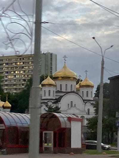 The image depicts an outdoor scene dominated by a white building with multiple golden domes, characteristic of an Orthodox church. The building's architecture features arched windows and decorative elements. In the foreground, a street light pole with wires stretches across the frame, obscuring parts of the view. A modern building with numerous windows, reflecting the light, stands behind the church. Trees and other greenery are visible in the background, suggesting an urban or suburban setting. The sky is overcast, indicating a cloudy or possibly rainy day. The overall impression is that of a community gathering place or a religious site within a developed area. The image appears to be taken during daylight hours, likely late afternoon or evening given the lighting conditions. No people are clearly visible, but the presence of a building suggests it is a public space. There is no visible text in the image.