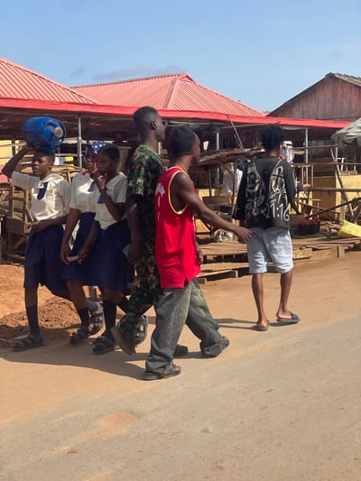 The image shows a group of people standing on a street in Ikoga, Nigeria. There are five people in the image, one of whom is carrying a blue gas tank on their head. The others are dressed in a variety of clothing, including a red tank top, a black and white patterned shirt, and school uniforms. The background includes buildings and other structures, suggesting a commercial or residential area. The street appears to be paved or partially paved.