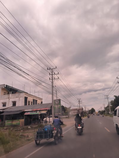The image presents an active street scene in Payameuneng, Indonesia, captured on an overcast day with a sky full of grey clouds. The perspective appears to be from a vehicle moving along a paved road, looking forward.
In the foreground, a man on a motorcycle is seen from behind, pulling a sidecar laden with several large blue water jugs. To his right, another motorcycle carries two people, with the passenger wearing a dark head covering. Further down the road, additional motorcycles and a white van are visible, suggesting moderate traffic.
Along the left side of the road, there are several buildings, including a multi-story structure and a line of smaller, open-fronted shops or stalls with corrugated metal roofs. Prominent signs above these shops display text such as "BU SIE KAMENG APADIN" and "BUSIEK MENG APA IN," indicating local businesses, possibly eateries. Numerous dense power lines crisscross the sky, supported by a series of utility poles that line both sides of the street, dominating the upper portion of the frame. Visible license plate numbers include "BL 6739 ZAT" on one of the motorcycles and a partial "BG5501日" on another.
The overall scene depicts a slice of daily life, characterized by local commerce, typical street infrastructure, and common modes of transport in a busy urban or semi-urban setting.
