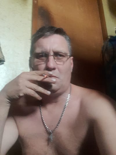A shirtless man with glasses is holding a lit cigarette to his lips. He is wearing a silver chain around his neck. The background appears to be indoors with wooden paneling. No specific location in Volokonovka, Russia can be determined from the image.