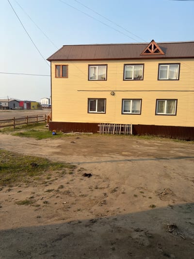 The image depicts a two-story, light yellow building with a brown metal roof, located in Verkhnevilyuysk, Russia. The building has several windows with white frames, some of which have curtains visible. A triangular wooden detail is present on the gable end of the roof. In front of the building, there is a dirt path with patches of grass and some scattered debris, including what appears to be animal droppings. A low wooden fence runs along the edge of the property. In the background, other buildings and vehicles are visible, suggesting a semi-urban or rural setting. A small blue and yellow toy vehicle is on the grass near the building. Power lines are visible in the sky. The lighting suggests it is either early morning or late afternoon.