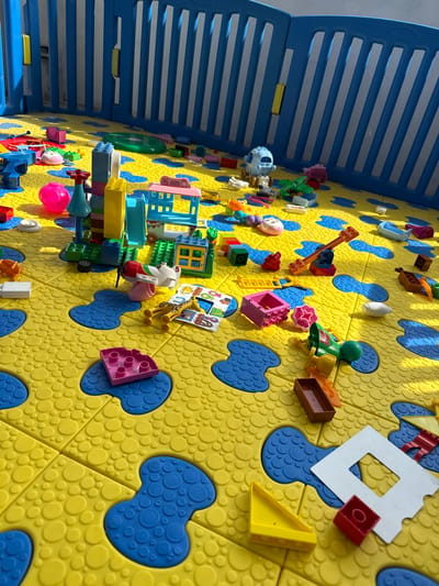 The image depicts a child's play area. A blue and white plastic playpen encloses a section of interlocking yellow and blue foam floor tiles. Numerous plastic toy blocks, including recognizable LEGO bricks, are scattered across the floor, some assembled into rudimentary structures. The lighting suggests an indoor setting with natural light. No people are visible. There is no information about Синьково, Russia in the image.