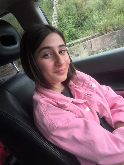 A young woman with dark hair and fair skin is sitting in the backseat of a car, with her left shoulder towards the viewer. She is wearing a pink denim jacket over a dark shirt. Her head is tilted slightly to her right, and she is looking towards the camera with a soft smile. Her makeup includes blush on her cheeks and lip gloss. The interior of the car is visible, with black leather seats and a headrest. Outside the car window, there is lush green vegetation and a concrete barrier, suggestive of a rural or semi-rural setting, possibly on the outskirts of Ribnovo, Bulgaria. The lighting appears to be natural daylight, casting a soft glow on the scene.