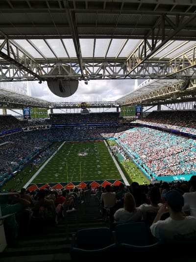 An elevated, wide-angle shot captures the interior of Hard Rock Stadium in Miami Gardens, United States, during a daytime American football game. The perspective is from an upper deck seat, looking down onto the expansive green football field where players are visible, albeit indistinctly, suggesting an ongoing match.
The upper portion of the image is dominated by the stadium's robust metal truss structure and translucent roof canopy, which provides shade to sections of the stands. A large, circular, dark object, possibly part of the stadium's sound system or central scoreboard, hangs suspended from this framework. The sky, partially visible above the stadium's structure, shows a mix of clouds.
The stadium's seating bowls are largely filled with spectators. The stands are sectioned, with some areas featuring teal and white seating, indicative of team colors. Two prominent video screens are visible on the far side of the stadium. The screen on the left displays a "BACARDÍ" logo and "MIA FINS" (referring to the Miami Dolphins), while the main screen shows game-related graphics including "J-1" and "J-2". The "Hard Rock" stadium name is prominently featured on a structure below the main video screen. The foreground spectators are mostly seen from behind, sitting in darker, shaded seats, observing the action on the brightly lit field.