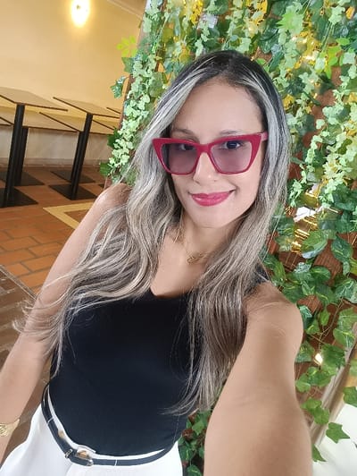 A woman with highlighted long blonde hair and red-framed sunglasses smiles at the camera. She is wearing a black tank top and white pants with a black and gold belt. The background includes a brick floor, tables, and a wall covered in green vines. She is wearing gold jewelry. The lighting is bright.
