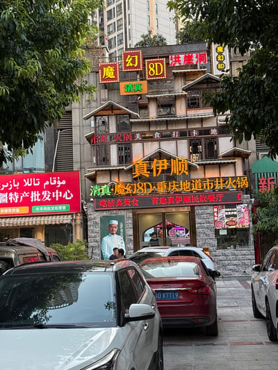 This image, taken in Longxi Subdistrict, China, depicts a bustling street scene with a focus on a multi-story building adorned with numerous illuminated signs, predominantly in Chinese characters. The building appears to be a restaurant or a commercial establishment, indicated by the signage "真伊顺" (Zhen Yi Shun), "吃重庆火锅" (Eat Chongqing Hotpot), and "新疆营养馕" (Xinjiang Nutritious Naan). There are also signs advertising "8D" experiences, suggesting some form of entertainment or unique offering.

In the foreground, several cars are parked along the sidewalk, with a prominent red car featuring a license plate that reads "D-X7719". A large red banner on the building displays text in both Chinese and what appears to be Arabic script, with Chinese characters translating to "Xinjiang Specialty Wholesale Center" and mentioning "beef and lamb wholesale and retail". A framed picture on the building shows a man in traditional attire, possibly a community leader or a representation of the restaurant's ethnic focus.

Lush green trees partially obscure the building, suggesting a temperate climate and possibly a daytime setting, although the bright neon signs create a vibrant, almost nighttime atmosphere. The architecture of the building is varied, with a mix of stone facades and wooden decorative elements, giving it a somewhat traditional yet commercial appearance. The overall impression is one of a lively urban environment with a strong emphasis on food and commerce, particularly highlighting Xinjiang cuisine and products.