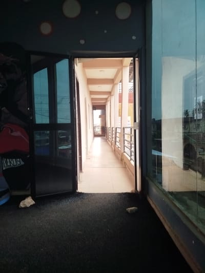 The image shows a long, covered walkway with an open-air corridor on the right. The walkway is lined with pillars and a metal railing, and the floor is tiled. On the left, there are large glass doors that reflect a mural of a boxing glove and the word "EVERLAST". The walkway leads into the distance, with buildings visible at the end. The time of day appears to be daytime, possibly late afternoon, given the lighting. The weather is unclear, but the reflections on the glass suggest it might be sunny. The scene is likely a public building or institution in Uselu, Nigeria, perhaps a school or a sports complex, given the boxing mural. There is no specific activity or event taking place, it appears to be an empty walkway.