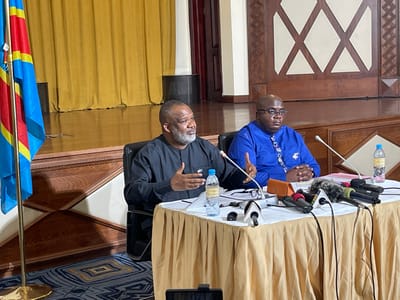 The image captures an indoor event, likely a press conference, featuring two men seated behind a draped table in the city of Nyayumba, Rwanda. The man on the left, with a grey beard and wearing a black long-sleeved shirt, is actively speaking, gesturing with both hands. The man on the right, bald and wearing glasses, is dressed in a blue patterned long-sleeved shirt, with his hands clasped in front of him.
The table is covered with a gold-colored tablecloth and holds multiple microphones with various colored windscreens, two gooseneck microphones, two plastic water bottles, papers, and a brown pouch. To the left of the frame, two national flags of the Democratic Republic of Congo are displayed on poles. The background features a raised platform with a polished wooden floor, yellow curtains on the left side, and wooden wall paneling with an "X" motif on the right. The floor in the immediate foreground is covered with a patterned carpet featuring blue, yellow, and black geometric designs.