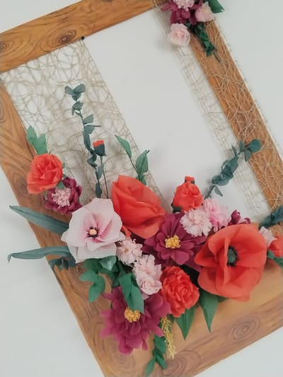 A close-up, eye-level, outdoor shot captures a decorative floral arrangement on a wooden frame, with a plain white background, presumably in Akberdino, Russia. The frame, made of light-colored wood with a visible grain pattern, is diagonally positioned. A net-like twine material is stretched across the opening of the frame. A vibrant bouquet of artificial flowers, primarily in shades of red, orange, pink, and burgundy, is attached to the lower portion of the frame. These flowers, appearing to be made of paper or tissue, include large poppy-like blooms, roses, and smaller multi-petaled flowers. Green paper leaves and stems accent the floral arrangement. The lighting is bright and even, casting no harsh shadows. The scene is static, with no people or animals present.