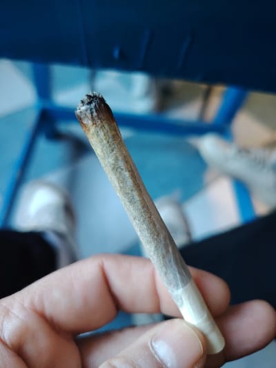 A hand holds a partially smoked rolled cigarette or marijuana joint, lit at the end. The joint has a tan color and is wrapped in thin white paper, tapering slightly towards the unlit end. Background features include a blue object, presumably a piece of furniture, and indistinct shapes that could be shoes and clothing. The overall scene suggests a casual, informal setting.