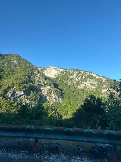 The image shows a mountain landscape under a clear blue sky. The mountains are covered with green trees and rocky outcrops. In the foreground, there is a metal guardrail with green spray paint markings and some gravel. The scene appears to be a roadside view, possibly in a mountainous region like the French Jura, given the mention of Septmoncel les Molunes, France. There are no people or animals visible in the image. The lighting suggests it is daytime, possibly late afternoon due to the angle of the shadows.