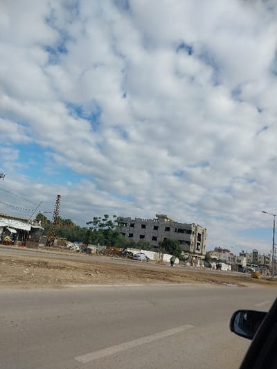 An outdoor, wide-angle street scene in a-Zawayda, Palestinian Territory, captured under a partially cloudy sky. The foreground features a two-lane asphalt road with a faded white lane marker, observed from the interior of a vehicle, evidenced by a dark side mirror frame in the bottom right. Beyond the road lies an unpaved, uneven dirt shoulder or verge containing scattered debris and areas of disturbed soil. To the left, several small, possibly temporary, structures with corrugated metal roofs are visible. Further right, a tall, red and white striped pole stands upright, resembling a barrier or gate. The central focus is a large, multi-story concrete building, approximately five floors tall, appearing either unfinished or damaged, characterized by numerous empty rectangular window openings and an unrendered facade. A white vehicle is partially obscured in the dirt in front of this structure. One individual, dressed in dark clothing, walks from right to left on the dirt path in front of the large building. Sparse green trees and bushes are situated between the road and the buildings. The background reveals additional residential or commercial structures, some appearing incomplete, and a street lamp pole on the far right. The sky is filled with numerous white cumulus clouds against patches of blue.