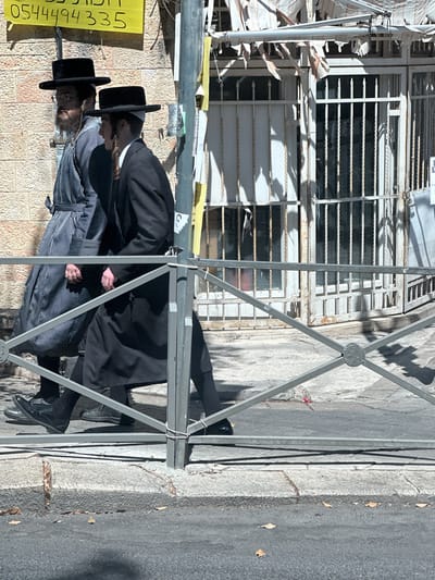 The image depicts two men, dressed in traditional Haredi Jewish attire, walking along a street in al-Eizariya, Palestinian Territories. Both men are wearing black hats and dark coats. One man is wearing a striped coat, while the other is in a solid black coat. They appear to be walking side-by-side, engaged in conversation or simply walking together. The setting is an urban environment with a building facade featuring barred windows and some scaffolding or construction materials. A yellow sign with Arabic text and numbers is visible on the left side of the image, which reads "054494335". The sun appears to be shining, casting shadows on the ground, suggesting it is daytime. The overall mood seems neutral and ordinary, depicting a moment of daily life in the location.