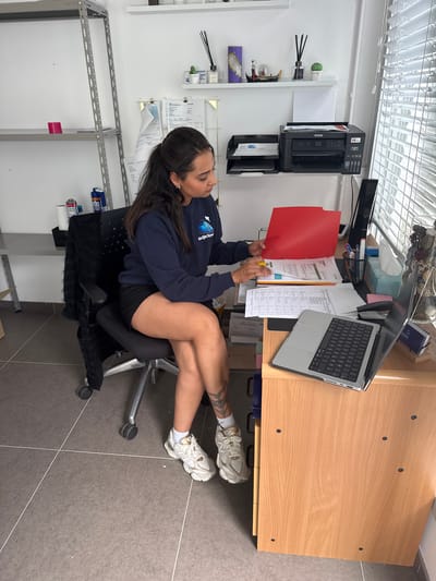 A woman sits at a desk in an office, sorting through papers. She is wearing a navy blue sweatshirt and black shorts, with white and gold sneakers. A laptop is open on the desk, and a red folder is being held by the woman. The office appears to be located in Maldegem, Belgium.
