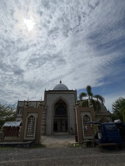 The image depicts the exterior of a mosque in Kruengmate, Indonesia, under a cloudy sky with the sun partially visible. The mosque features a prominent dome and is constructed with stone-like tiles. In front of the mosque, there are decorative fences and a blue vehicle that appears to be a small truck or van, possibly used as a vendor's stall with items displayed on its side. A palm tree is visible to the right of the mosque. The ground in the foreground is covered with gravel.