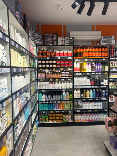 This indoor shot depicts a section of a store, likely in Аксукент, Kazakhstan, focusing on shelves filled with various hair and skincare products. The shelves are predominantly stocked with shampoos, conditioners, and cosmetic items. Brands such as "LAGOM", "ARVER", "LISON", "café mimi", "MAC", "HAARTEC", "PROMOZE", "fino", "SOKAN", "THERAS", "POWER", "IR PEELING", and "cansong" are visible. The products are arranged in neat rows within the shelving units, with some shelves displaying testers. The lighting fixtures, consisting of multiple spotlights, are mounted on the ceiling, highlighting the products on display. The store's color scheme includes white, grey, black, and various product packaging colors. The floor appears to be tiled. An air conditioning unit is mounted on the wall, next to the shelves. A pink bag sits near the top right corner of the image. The photograph appears to have been taken from a medium-high angle, giving a comprehensive view of the product displays.