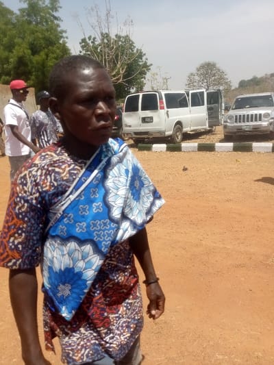 An outdoor scene in Kagoma, Nigeria, depicts an unpaved, reddish-brown dirt ground under clear daylight. In the immediate foreground, a dark-skinned individual with short, dark hair wears a multicolored patterned garment featuring orange, brown, and blue tones, complemented by a blue and white patterned fabric draped across their left shoulder and chest. A dark bracelet is visible on their right wrist. The individual faces slightly right of the viewer, exhibiting a neutral expression.
Behind this primary subject, two other individuals are partially visible on the left. One person, wearing a white t-shirt and a red baseball cap, stands to the left. Further back, another person wears a patterned shirt.
In the midground, beyond a low concrete curb painted with alternating black and white sections, two vehicles are parked on a paved or hardened surface. A white panel van, with visible Nigerian license plate "KJA 93XJ" on its rear, has both rear doors open. To its right, a silver SUV, possibly a Jeep, is parked, displaying a partially visible license plate beginning with "LSD-947".
The background features sparse green trees and shrubs under a light blue sky. Distant hills or elevated terrain are visible on the far right horizon. The overall environment appears dry and open.