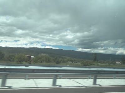 The image depicts a view from a highway near Agarak, Armenia. A modern concrete and metal guardrail runs across the foreground. Beyond the guardrail is a green field with scattered trees and bushes. In the distance, a long mountain range stretches across the horizon, with some snow visible on the peaks. The sky is overcast with heavy gray clouds, with a sliver of blue sky visible near the mountain range. There is a vapor trail in the sky. A red-roofed house is partially obscured by trees.
