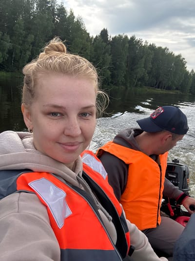 Two individuals are on a boat, the woman in the foreground is taking a selfie. She has blonde hair pulled up in a messy bun and is wearing a beige hoodie and an orange life vest. The man behind her is wearing a dark baseball cap and an orange life vest, and is operating the boat's outboard motor. The boat is moving on a body of water, with a dense forest lining the shore in the background. The sky is overcast. The scene appears to be in a natural environment, likely a river or lake. The location is Anteplevo (Okhotinskoe S/P), Russia.