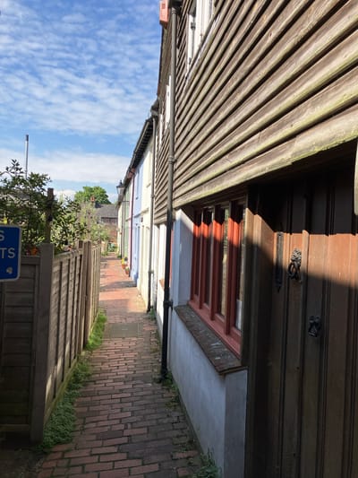 The image presents a narrow alleyway in Lewes, United Kingdom, paved with red bricks. Wooden fences and building façades border the path on either side. The buildings exhibit various colors, including white, blue, and pink. At the end of the alley is green foliage. The sky above is blue with scattered white clouds. A wooden structure looms on the left, with horizontal planks. A wooden door is to the right, adorned with metal fittings and a window below. A black pipe runs along the wall. A blue sign is visible near the bottom of the image.