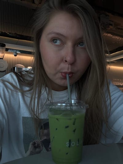A young woman with blonde hair and blue eyes is holding a green drink. She has a nose ring and is drinking from the cup with a straw. The drink is a light green color with ice cubes. The brand name "SOLA" is printed on the cup. The woman is wearing a white t-shirt. The background is blurred, but it appears to be a cafe or restaurant. The picture is angled.