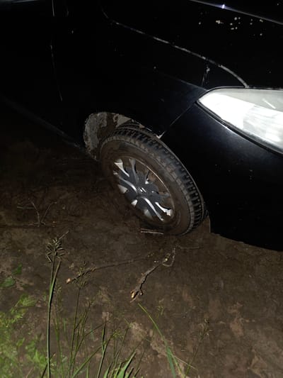 The image shows a black car stuck in mud. The front passenger-side wheel of the car is partially submerged in thick, wet mud. The car's fender and headlight are visible, as is the tire and rim of the stuck wheel. The surrounding environment is muddy ground with some sparse vegetation and twigs scattered on the surface. The scene appears to be outdoors, possibly in a rural or undeveloped area, with no clear signs of city infrastructure. The lighting suggests it may be nighttime or a very dimly lit area. The specific location in the city of Tagashevo, Russia, is not discernible from the visual information in the image alone.