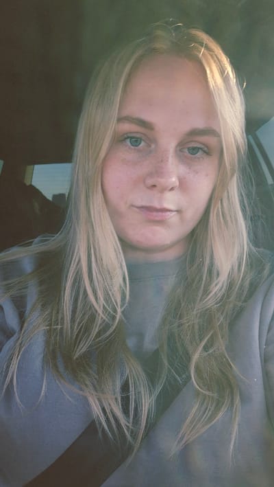 The image is a close-up, head-and-shoulders selfie of a young woman with blonde hair and blue eyes. She has freckles across her nose and cheeks. She is wearing a grey sweatshirt and a black seatbelt is visible across her chest. The background is blurred, but appears to be the interior of a vehicle. The lighting suggests it is daytime. The location is identified as Razeni, Moldova.