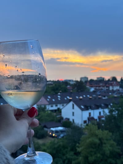 A person's hand with bright red nail polish holds a wine glass filled with white wine, in front of a sunset sky over a residential area. The glass has condensation droplets on its surface. The background shows rooftops of houses, some with dormer windows, and trees. The sky displays hues of blue and orange from the setting sun. The scene appears to be from Sindelfingen, Germany, based on the architecture and typical residential appearance.