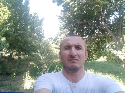 A man in a white t-shirt is looking directly at the camera, with a slight grimace. He is in an outdoor setting with trees and greenery in the background. The man appears to be in his 40s, with short, dark hair and a stubbly beard. The lighting suggests it is daytime, possibly late morning or early afternoon, given the brightness and the shadows cast by the trees. The environment looks like a park or a rural area. There is no specific indication of it being in Karagana, Uzbekistan, within the image itself.