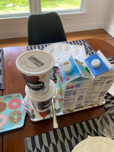 A wooden table in a kitchen or dining area, at a residence located in Rye, New York, coordinates (40.952, -73.708). There are two containers of "Liberte Greek" yogurt, one open and one closed. Next to the containers of yogurt is a small, rectangular, multi-colored serving tray. On the tray, from left to right are: a ceramic plate with a blue background, a white design, and a colorful donut pattern; a stainless steel spoon; a white and blue rooster design on the tray; and two 100% juice boxes, Dole Orange-Mango, with labels and nutritional information. There is a white napkin to the right of the yogurt. A black chair is visible to the left of the table and a window with a view of green trees.