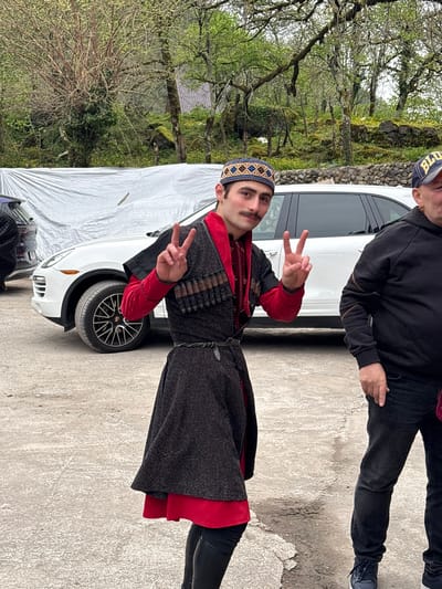 A man dressed in a traditional Georgian Chokha costume and a hat stands with a car in the city of Kveda Chkhutuneti, Georgia. He is making a peace sign with both hands. The background includes trees, a building, and a stone wall. A second man is partially visible, wearing a black jacket and jeans. The ground appears to be concrete.