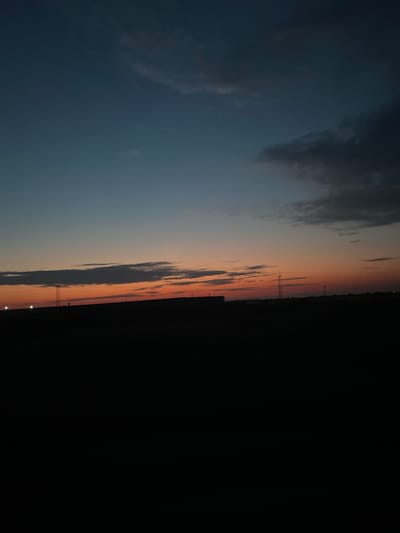 The image depicts a twilight sky over a dark, silhouetted landscape. The sky transitions from a deep blue at the top to shades of orange and red near the horizon. Clouds are visible, with some appearing darker and heavier than others. The lower portion of the image is entirely black, obscuring any details of the ground, structures, or any potential human presence. No specific identifiable landmarks of Il'inka, Russia are visible due to the darkness and the nature of the scene. The overall impression is one of a barren or undeveloped area at dusk.