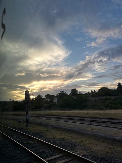 The image captures a sunset view from a train, looking out over a railway track and the surrounding landscape of Alès, France. The sky is filled with dramatic clouds, ranging from dark grey to bright white and tinged with the warm hues of the setting sun. Sunlight breaks through the clouds in a bright, horizontal band. In the foreground, a set of train tracks runs parallel to the viewer, disappearing into the distance. To the left of the tracks, a vintage signal post stands tall, its structure partly obscured by shadow and the reflection from the train window. Beyond the tracks, a grassy embankment is dotted with trees and houses, some with red-tiled roofs, suggesting a suburban or semi-rural setting on the outskirts of Alès. The overall impression is one of a peaceful, late afternoon or early evening scene.