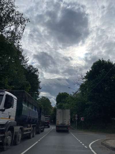 The image shows a road in Semyonovskoye, Russia, under a cloudy sky. Several trucks are on the road, including a white one in the foreground. Trees line both sides of the road. There are traffic signs visible near a curve in the road further ahead. The sun shines through the clouds in the sky.