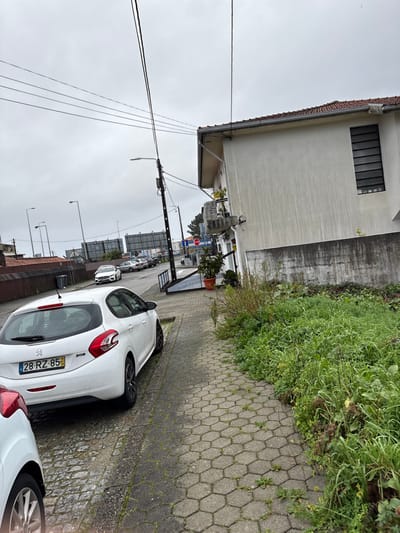 The scene depicts an urban street on an overcast day in Santa Maria da Feira, Portugal. In the immediate foreground, the right side shows a hexagonal paver sidewalk with weeds growing through cracks, bordering an unkempt strip of dense, green vegetation. The left foreground shows the rear of a white Peugeot 208 hatchback (license plate 28-RZ-85) parked on what appears to be a cobblestone or roughly paved street. The front portion of a second white vehicle is partially visible on the far left.

To the right of the parked Peugeot, a dark grey utility pole with an attached street lamp stands beside the sidewalk. Overhead, multiple black utility lines traverse the frame horizontally and diagonally. Further along the sidewalk, a ramp made of dark metal scaffolding leads up to the ground-floor entrance of a two-story, light-colored stucco building. The building features a red-tiled roof, a small balcony on the second floor with a potted plant, and a horizontally barred window on its right facade. Staining is visible on the building's exterior walls. Another large potted plant sits near the building's entrance. A red octagonal "STOP" sign is visible near the entrance.

In the mid-ground, the street continues with several other cars parked parallel to the curb. A dark grey rubbish bin is visible on the left side of the street. The background features additional streetlights, large billboard-like structures without visible advertisements, and more distant buildings under the uniform grey sky. No individuals are present in the image.