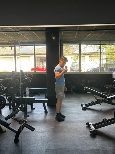 A man in a light blue shirt and gray shorts is looking at his phone while standing in a gym. The gym has weightlifting equipment and large windows. Outside the windows are views of houses and cars. The scene is located in Jestetten, Germany.