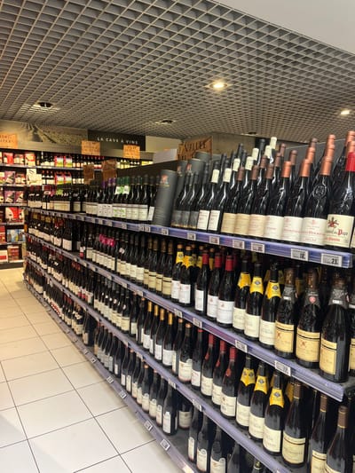 The image shows a wine aisle in a grocery store located at latitude 49.283 and longitude 3.999 in Saint-Brice-Courcelles, France. There are rows of wine bottles on metal shelves, organized by brand and type. The shelves are labeled with prices and information about the wines. The store has a white tiled floor and a white ceiling with black grid-style panels. The aisle is well lit. The sign above the shelves reads “La Cave à Vins” which translates to "The Wine Cellar". There are no visible people in the image. The wine bottles are all full and appear to be in good condition. The bottles are arranged in a neat and orderly fashion. There is no visible damage to the shelves or the bottles.